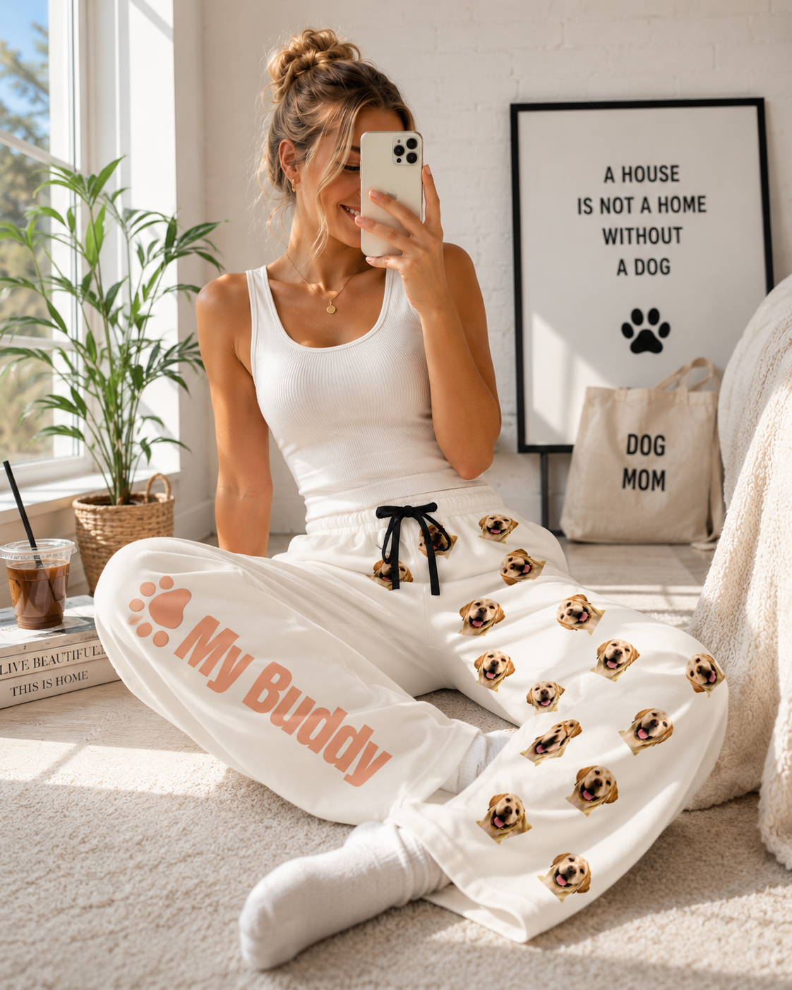 A woman sitting on a carpeted floor in a bright room, taking a mirror selfie while wearing a white tank top and white sweatpants with gold and black dog face prints and the text 'My Buddy'.